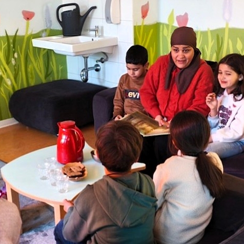 Eine Mutter liest Kindern auf Arabisch vor. Die Kinder sitzen aufmerksam auf einem Sofa in gemütlicher Runde.