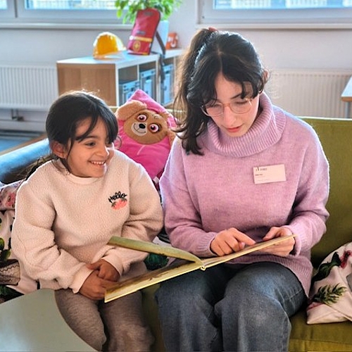 Eine Praktikantin liest einem Kind auf dem Sofa eine Geschichte vor. Beide wirken entspannt und fröhlich.