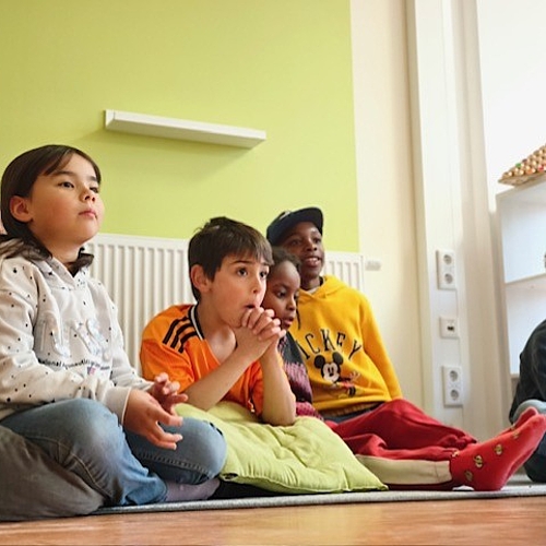 Eine Gruppe Kinder verschiedener Herkunft hört konzentriert zu, während im Hintergrund helles Tageslicht den Raum erfüllt.