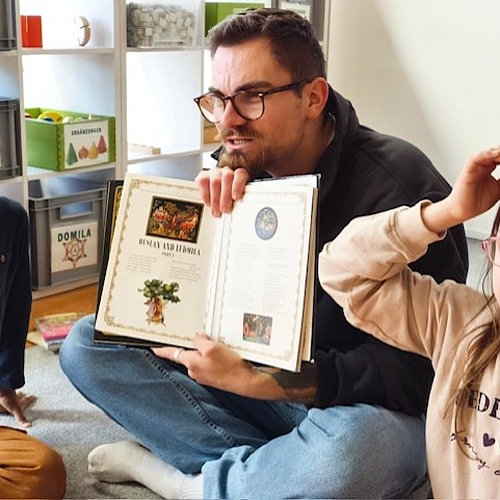 Tom liest mit sichtbarem Engagement eine englische Geschichte vor, Kinder sitzen dicht bei ihm auf dem Teppich.