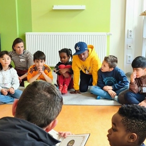Eine Gruppe Kinder sitzt im Halbkreis und hört aufmerksam einem Mann beim Vorlesen auf Englisch zu.