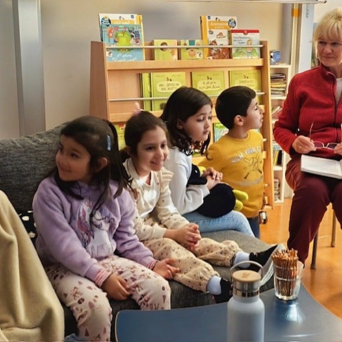 Eine Erzieherin liest einer Gruppe Kinder auf Deutsch vor. Die Kinder sitzen nebeneinander auf einem Sofa und hören gespannt zu.