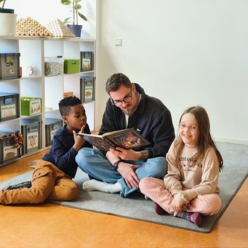 Tom sitzt mit zwei Kindern auf dem Teppich und liest ein englisches Buch, ein Kind schaut interessiert, das andere lächelt.